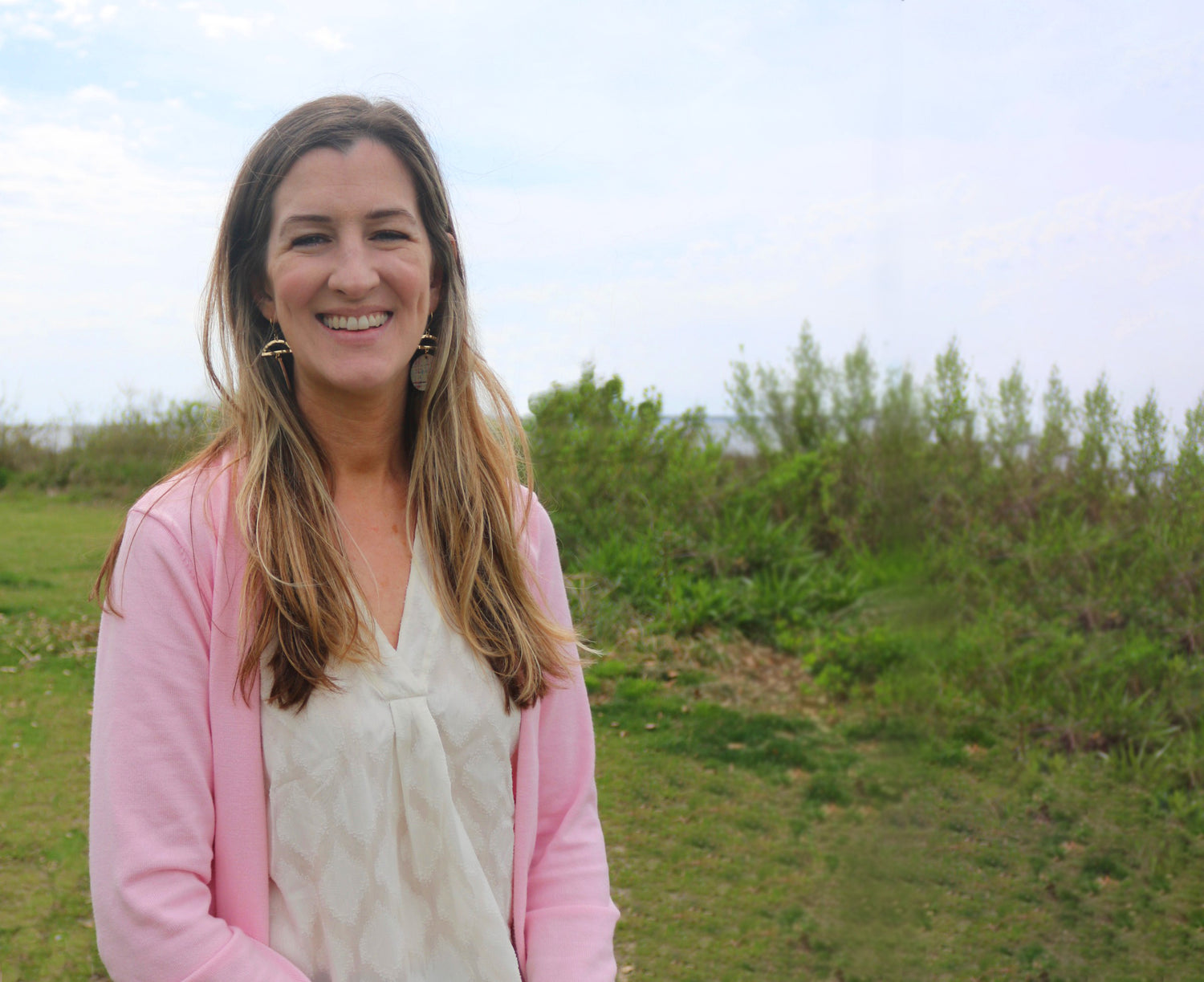 Woman in a pink cardigan standing outdoors with greenery in the background